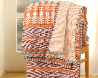 May include: A folded orange and white patterned quilt draped over a wooden chair. The quilt features floral and chevron designs in shades of orange, brown, and white. The reverse side has vertical stripes.