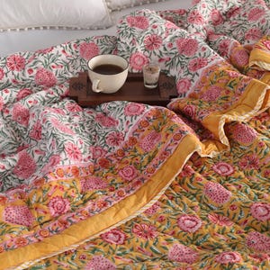 Könnte beinhalten: Eine florale Steppdecke mit rosa Blumen auf weißem Grund, akzentuiert mit gelben Rändern. Ein Holztablett hält eine Tasse Tee und ein kleines Glas. Die Steppdecke liegt auf einem Bett mit weißen Kissen.