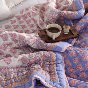Peut inclure: Une couverture matelassée à motifs floraux roses et blancs, avec une bordure bleue. Un plateau en bois contient une tasse de thé et une petite bougie. La couverture est sur un lit blanc.