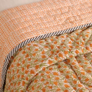 May include: A quilted bedspread with a floral pattern in orange and green on a white background. The top section has an orange and white arrow pattern. A black and white striped border separates the two patterns.