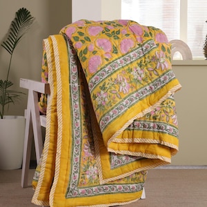 May include: A yellow and pink floral patterned quilt with a yellow and white striped border. The quilt is folded and draped over a white wooden chair.