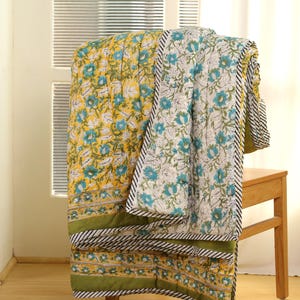May include: A floral patterned quilt draped over a wooden chair. The quilt features a yellow background with blue and white flowers. The border is green with black and white stripes. The quilt is made of soft, comfortable material.
