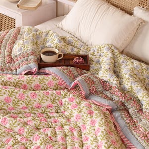 Puede incluir: Una cama cubierta con un edredón floral en rosa y verde. Una bandeja de madera contiene una taza de café y una flor decorativa. Almohadas blancas y un cabecero tejido completan la acogedora escena del dormitorio.