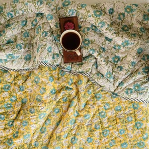 Puede incluir: Un edred&oacute;n floral reversible con un dise&ntilde;o floral blanco y turquesa en un lado y un estampado floral amarillo y turquesa en el otro. Una bandeja de madera con una taza de caf&eacute; descansa sobre el edred&oacute;n.