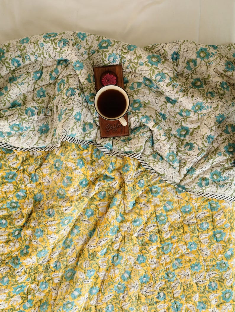 Puede incluir: Un edred&oacute;n floral reversible con un dise&ntilde;o floral blanco y turquesa en un lado y un estampado floral amarillo y turquesa en el otro. Una bandeja de madera con una taza de caf&eacute; descansa sobre el edred&oacute;n.