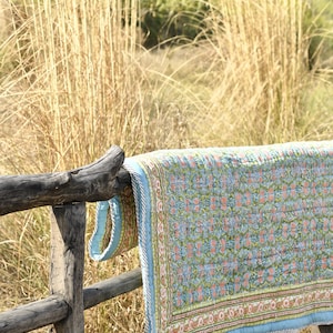 Może przedstawiać: Niebieska i zielona pikowana kołdra z kwiatowym wzorem jest przewieszona przez drewniane ogrodzenie. Kołdra ma jasnoniebieski brzeg z białym i zielonym wzorem kwiatowym.