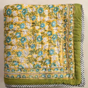 Puede incluir: Una manta acolchada plegada con un estampado floral. La manta presenta un fondo amarillo con dise&ntilde;os florales azules y blancos. El borde es verde con un borde a rayas blanco y negro. El edred&oacute;n est&aacute; hecho de un material acolchado suave.