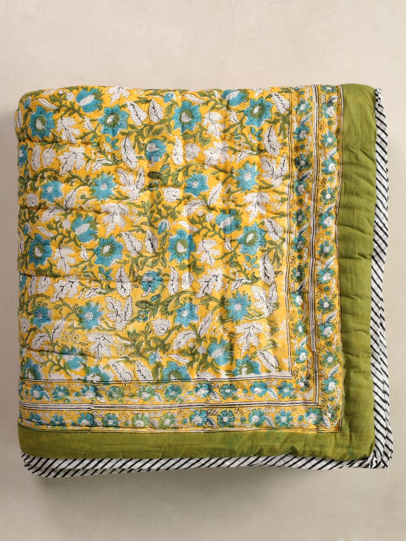 Puede incluir: Una manta acolchada plegada con un estampado floral. La manta presenta un fondo amarillo con dise&ntilde;os florales azules y blancos. El borde es verde con un borde a rayas blanco y negro. El edred&oacute;n est&aacute; hecho de un material acolchado suave.
