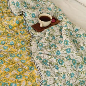 Puede incluir: Un edred&oacute;n reversible con un estampado floral en amarillo y blanco con detalles en azul. El edred&oacute;n tiene un borde a rayas blanco y negro. Una taza de t&eacute; est&aacute; sobre una bandeja de madera encima del edred&oacute;n.