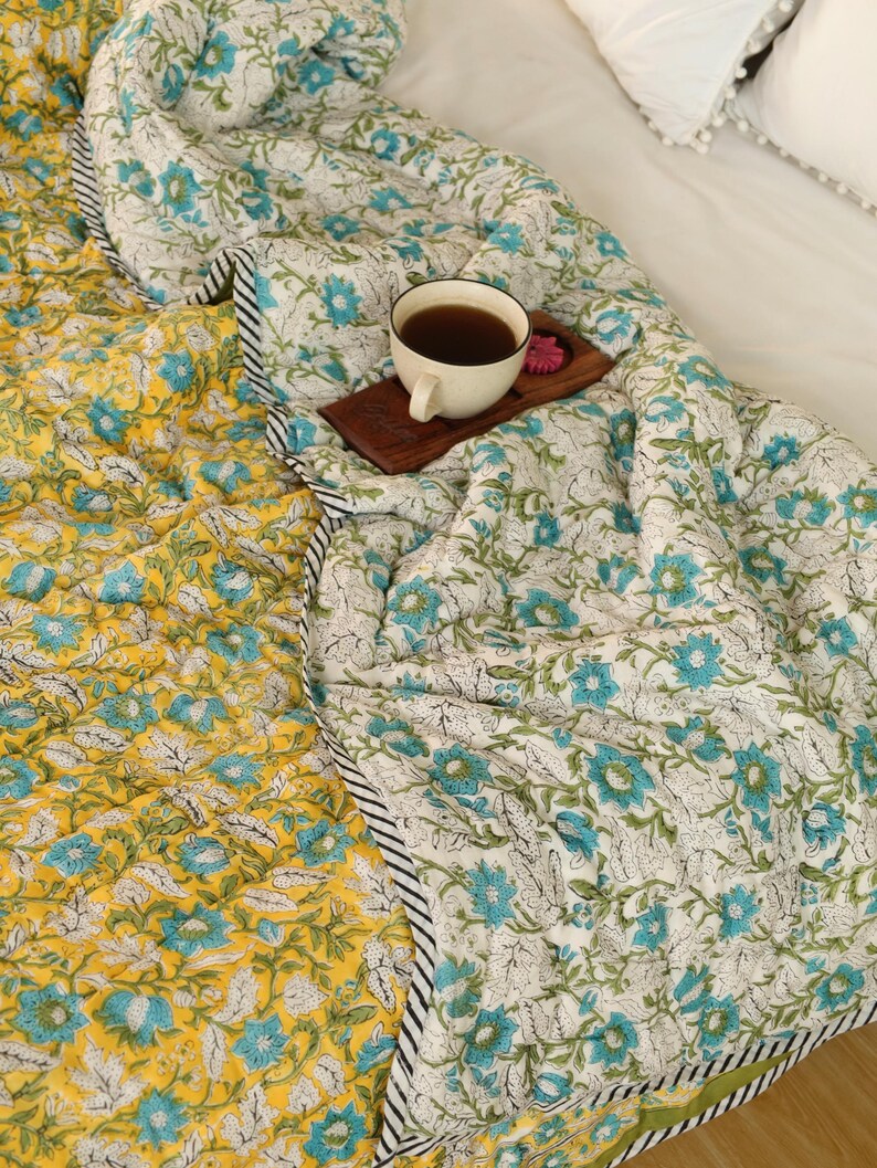 Puede incluir: Un edred&oacute;n reversible con un estampado floral en amarillo y blanco con detalles en azul. El edred&oacute;n tiene un borde a rayas blanco y negro. Una taza de t&eacute; est&aacute; sobre una bandeja de madera encima del edred&oacute;n.