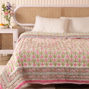Pode incluir: Uma cama branca com uma colcha estampada com flores rosa e verdes. A cama tem uma cabeceira de vime branca e uma mesa de cabeceira branca com uma gaveta de vime.