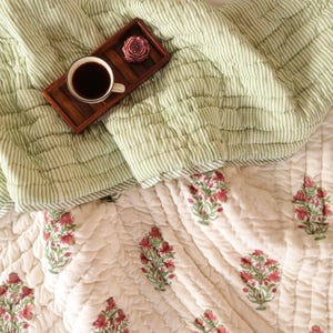 Peut inclure: Une courtepointe à rayures vertes et blanches avec un oreiller assorti. Un plateau en bois contient une tasse de café et une fleur décorative. Le bas de la courtepointe a un fond crème avec des motifs floraux rouges.