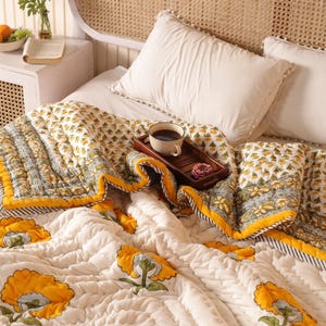 May include: A bed with a white headboard and a floral quilt. The quilt features large yellow flowers on a cream background, with a yellow border. A wooden tray with a cup of coffee and a rose sits on the quilt. White pillows complete the bedding.