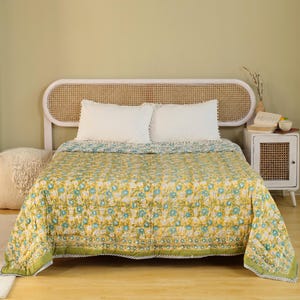 Puede incluir: Una colcha con estampado floral amarillo y azul sobre una cama con almohadas blancas. La colcha tiene un borde verde y est&aacute; colocada sobre una estructura de cama de madera. La habitaci&oacute;n tiene una paleta de colores neutros.