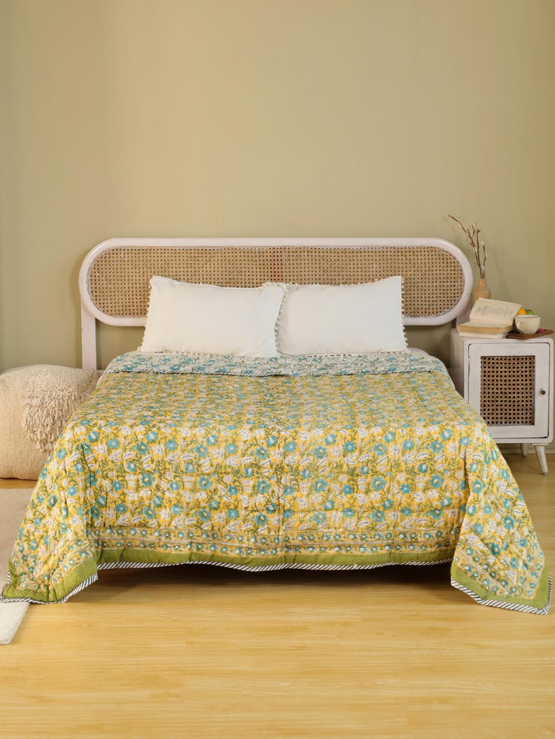 Puede incluir: Una colcha con estampado floral amarillo y azul sobre una cama con almohadas blancas. La colcha tiene un borde verde y est&aacute; colocada sobre una estructura de cama de madera. La habitaci&oacute;n tiene una paleta de colores neutros.