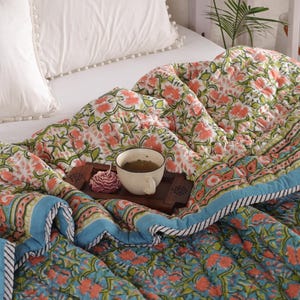 May include: A blue and green floral patterned quilt with a white border and a white pillow with a white pom-pom trim. A wooden tray with a cup of tea and a pink flower sits on top of the quilt.