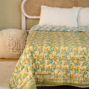 Puede incluir: Un edred&oacute;n floral amarillo con flores azules y blancas, borde verde y almohadas blancas sobre una cama. El edred&oacute;n tiene un patr&oacute;n repetitivo de flores y hojas. La cama tiene un cabecero tejido y un puf crema.