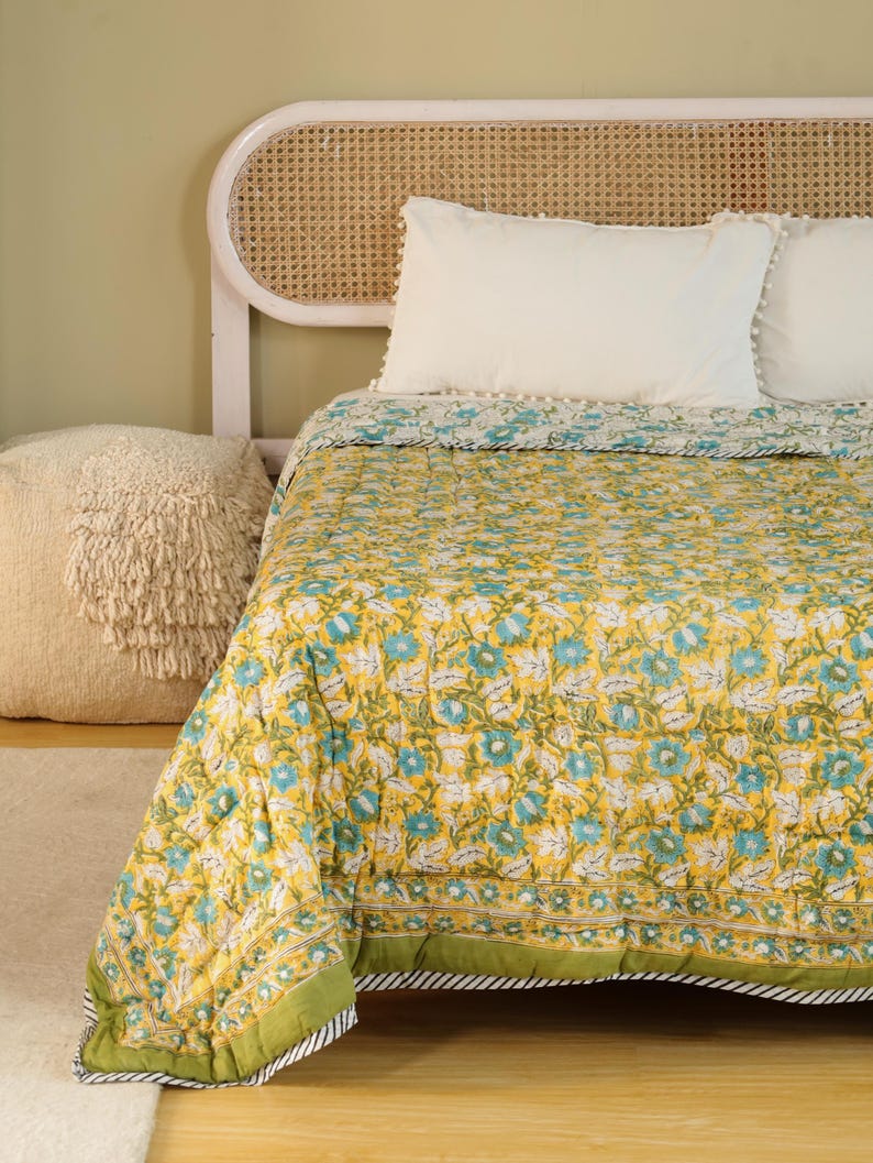 Puede incluir: Un edred&oacute;n floral amarillo con flores azules y blancas, borde verde y almohadas blancas sobre una cama. El edred&oacute;n tiene un patr&oacute;n repetitivo de flores y hojas. La cama tiene un cabecero tejido y un puf crema.