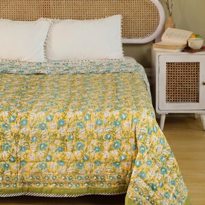 Puede incluir: Un edred&oacute;n amarillo floral con detalles azules y blancos, exhibido en una cama con almohadas blancas. El edred&oacute;n tiene un borde verde con un dise&ntilde;o a rayas. El cabecero es de madera clara.