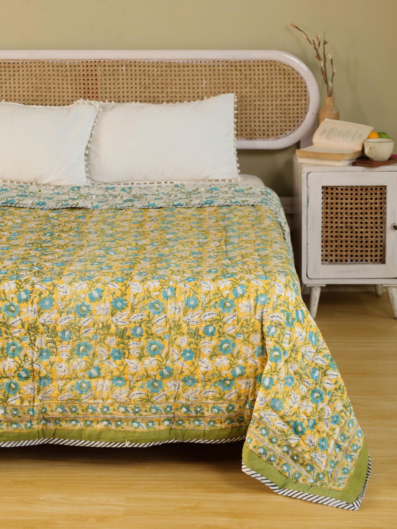 Puede incluir: Un edred&oacute;n amarillo floral con detalles azules y blancos, exhibido en una cama con almohadas blancas. El edred&oacute;n tiene un borde verde con un dise&ntilde;o a rayas. El cabecero es de madera clara.