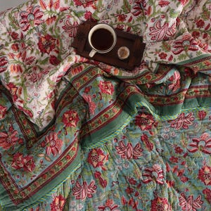 May include: A floral patterned quilt with a green and red border. The quilt is folded in half and a cup of coffee is sitting on top of it.