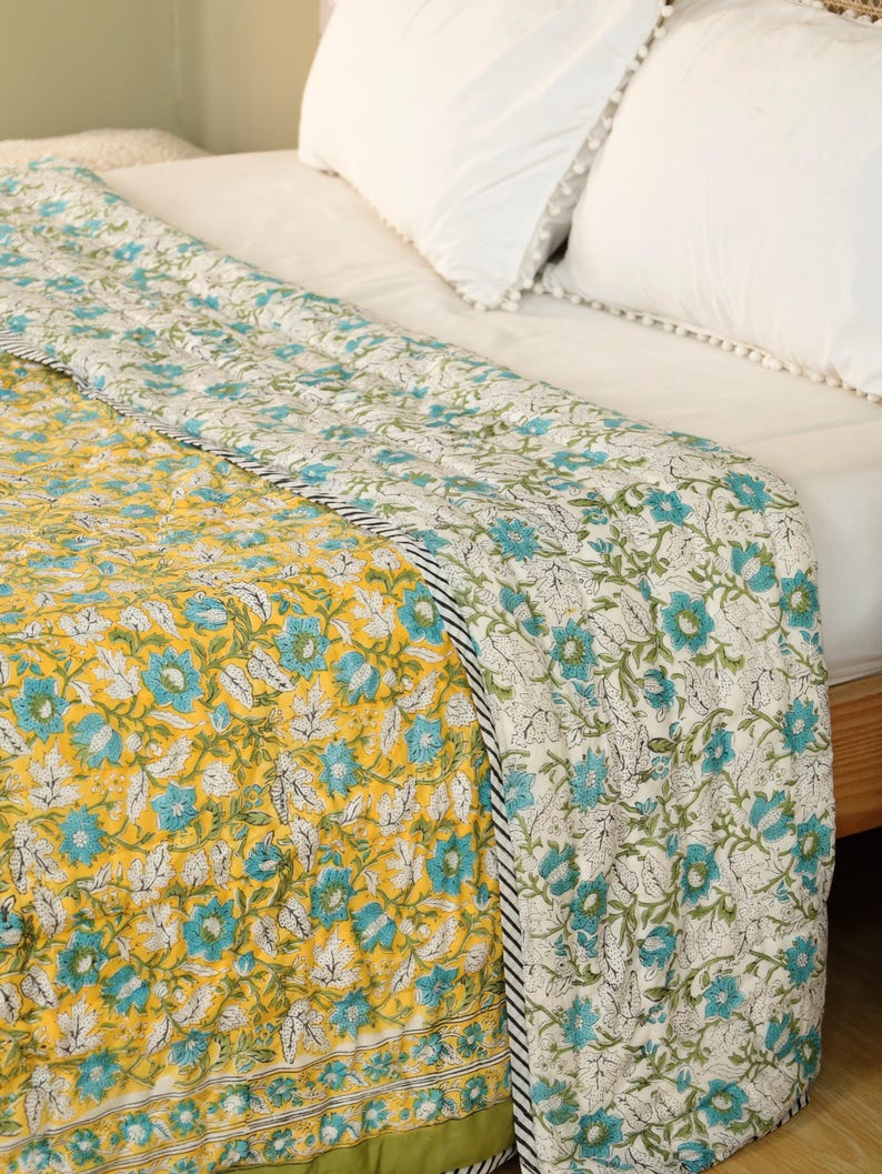Puede incluir: Un edred&oacute;n con estampado floral en tonos amarillos, turquesa y blanco. El edred&oacute;n presenta un patr&oacute;n repetido de flores y hojas. El borde es una raya en blanco y negro. El edred&oacute;n est&aacute; sobre una cama con almohadas blancas.