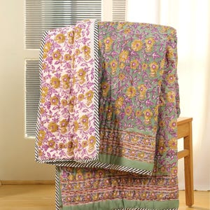 May include: Two floral patterned quilts, one in white with purple and gold flowers, the other in green with similar floral designs. Both quilts have a black and white striped border and are draped over a wooden stool.