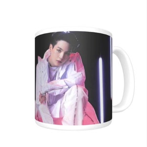 May include: White ceramic mug with a black background featuring a photo of a person wearing a white jacket and pants. The person is sitting on a pink surface with neon lights in the background.