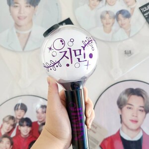 K&ouml;nnte beinhalten: Ein lila-schwarzer Lichtstick mit dem koreanischen Wort "Jimin" darauf. Der Lichtstick befindet sich in einer durchsichtigen Plastikkugel mit einem schwarzen Griff.