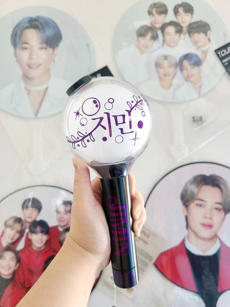 K&ouml;nnte beinhalten: Ein lila-schwarzer Lichtstick mit dem koreanischen Wort "Jimin" darauf. Der Lichtstick befindet sich in einer durchsichtigen Plastikkugel mit einem schwarzen Griff.