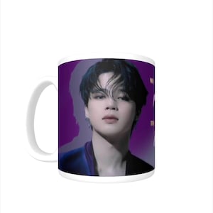 May include: White ceramic mug with a purple background featuring a portrait of a person with dark hair. The person is wearing a blue shirt and has a serious expression. The text "Mikrokosmos" is visible on the mug.