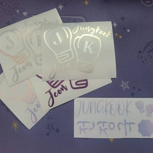 K&ouml;nnte beinhalten: Vier wei&szlig;e Vinyl-Aufkleber mit einem Boxhandschuh-Design. Die Handschuhe sind silbern umrandet und haben einen Buchstaben J oder K im Inneren. Die Aufkleber sagen "Jungkook" und "Jeon" in einer silbernen Schrift.