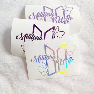 Può includere: Tre adesivi in ​​vinile bianco con finitura olografica. Gli adesivi presentano un design stilizzato di due linee verticali con una farfalla su ogni lato. Il testo "Military Wife" è scritto in un carattere corsivo sotto il design.