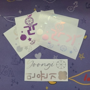 K&ouml;nnte beinhalten: F&uuml;nf wei&szlig;e Aufkleber mit verschiedenen Designs. Die Aufkleber zeigen eine lila Zitrusfrucht mit dem Text "SUDA" und dem koreanischen Wort "귤" auf einem Aufkleber, eine rosa Zitrusfrucht mit dem koreanischen Wort "귤" auf einem anderen Aufkleber und eine silberne Zitrusfrucht mit dem Text "Goongi" und dem koreanischen Wort "귤" auf einem dritten Aufkleber. Die restlichen beiden Aufkleber zeigen eine blaue Zitrusfrucht und ein wei&szlig;es Herz.