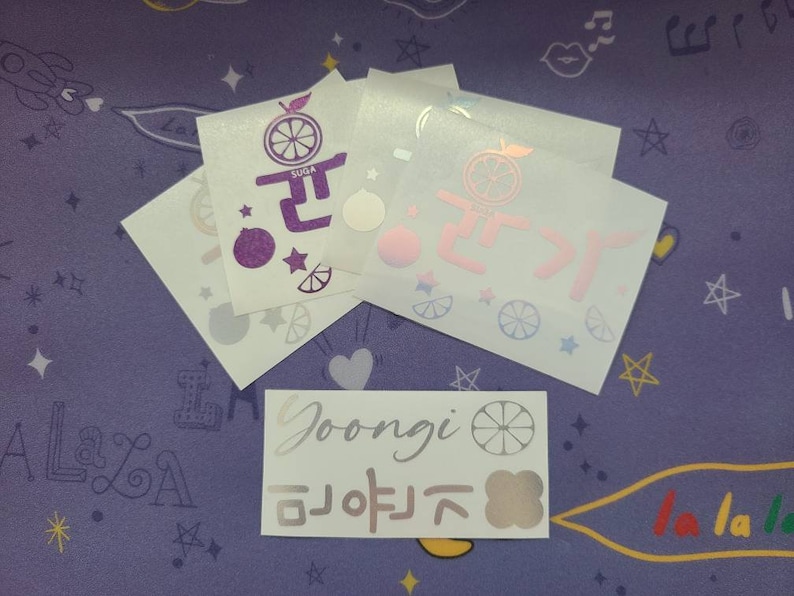 K&ouml;nnte beinhalten: F&uuml;nf wei&szlig;e Aufkleber mit verschiedenen Designs. Die Aufkleber zeigen eine lila Zitrusfrucht mit dem Text "SUDA" und dem koreanischen Wort "귤" auf einem Aufkleber, eine rosa Zitrusfrucht mit dem koreanischen Wort "귤" auf einem anderen Aufkleber und eine silberne Zitrusfrucht mit dem Text "Goongi" und dem koreanischen Wort "귤" auf einem dritten Aufkleber. Die restlichen beiden Aufkleber zeigen eine blaue Zitrusfrucht und ein wei&szlig;es Herz.