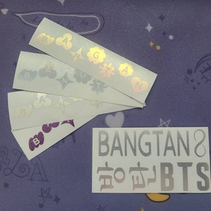 K&ouml;nnte beinhalten: Ein Satz von f&uuml;nf holografischen Aufklebern mit den Buchstaben "BANGTAN" und den koreanischen Schriftzeichen f&uuml;r "BTS" in silberner Schrift. Die Aufkleber befinden sich auf einem violetten Hintergrund mit wei&szlig;en Sternen.