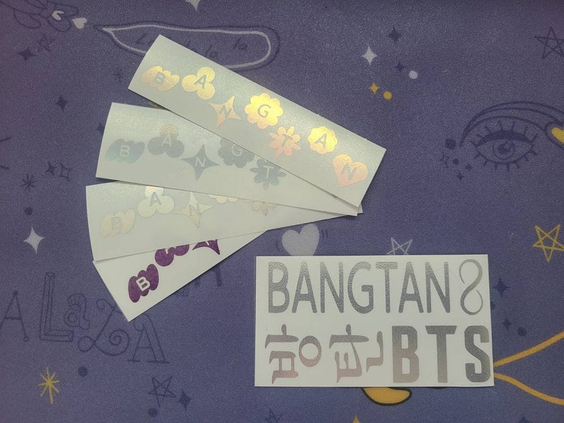 K&ouml;nnte beinhalten: Ein Satz von f&uuml;nf holografischen Aufklebern mit den Buchstaben "BANGTAN" und den koreanischen Schriftzeichen f&uuml;r "BTS" in silberner Schrift. Die Aufkleber befinden sich auf einem violetten Hintergrund mit wei&szlig;en Sternen.