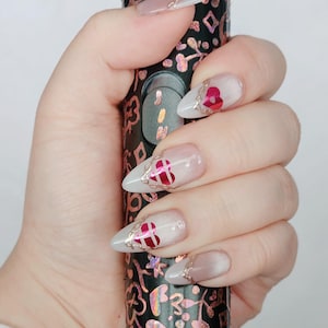 Puede incluir: Una mano con uñas largas y puntiagudas, manicuradas. Las uñas están pintadas con una base transparente, puntas blancas y diseños de corazón rojo. Las uñas están decoradas con detalles dorados. La mano sostiene un objeto negro con un patrón floral.