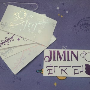 K&ouml;nnte beinhalten: F&uuml;nf wei&szlig;e Aufkleber mit violetten und silbernen Designs. Der obere Aufkleber tr&auml;gt das koreanische Wort "Jimin" in Silber. Der untere Aufkleber tr&auml;gt das Wort "Jimin" in violetten Buchstaben und koreanischen Text in Violett.