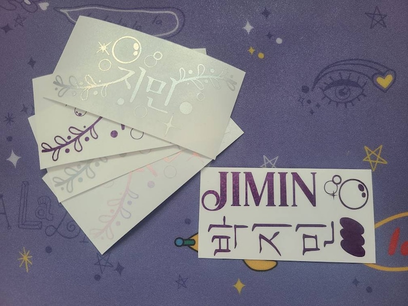 K&ouml;nnte beinhalten: F&uuml;nf wei&szlig;e Aufkleber mit violetten und silbernen Designs. Der obere Aufkleber tr&auml;gt das koreanische Wort "Jimin" in Silber. Der untere Aufkleber tr&auml;gt das Wort "Jimin" in violetten Buchstaben und koreanischen Text in Violett.