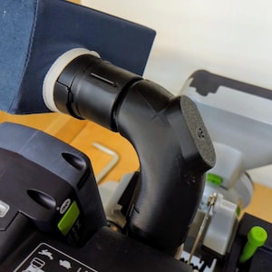 May include: A black plastic dust collection elbow attached to a power tool. The elbow connects to a dark blue fabric dust bag with a white ring. The power tool has black and gray components with green accents.