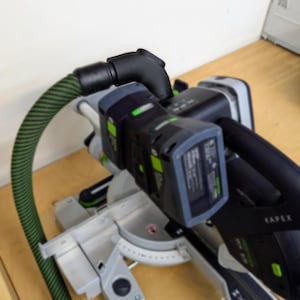 May include: A black and green power tool with a green hose attached to the side. The tool has a battery pack and a black handle. The tool is sitting on a wooden surface.