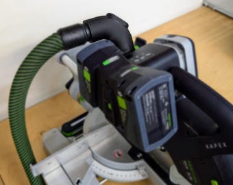 May include: A black and green power tool with a green hose attached to the side. The tool has a battery pack and a black handle. The tool is sitting on a wooden surface.