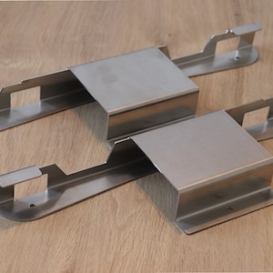 Op de afbeelding: Twee zilveren metalen beugels met uitsparingen en afgeronde randen. De beugels zijn ontworpen om aan een oppervlak te worden bevestigd en hebben een rechthoekige vorm.