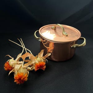 Könnte beinhalten: Ein kleiner Kupfertopf mit Deckel und Messinggriffen. Der Topf steht auf einer schwarzen Oberfläche neben einem kleinen Bündel getrockneter orangefarbener Blumen. Der Topf ist rund und hat eine glänzende Oberfläche.