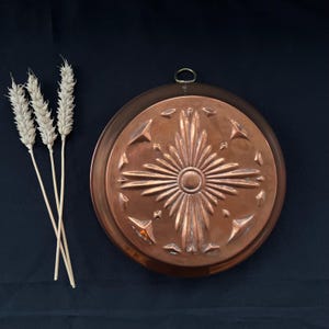 May include: A copper-colored, round decorative mold with a central sunburst design and a hanging loop. The mold is displayed on a dark background, with several stalks of wheat to the left.