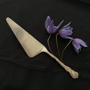 Puede incluir: Un servidor de pastel plateado con un mango decorativo, junto a tres flores moradas. El servidor tiene una hoja triangular y un diseño floral detallado en el mango. Las flores tienen pétalos delicados y tallos delgados.