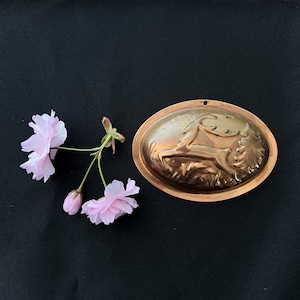 May include: An oval copper mold with a raised design of a deer leaping through tall grass. The mold has a small hole at the top for hanging. Pink cherry blossoms are arranged near the mold against a black background.
