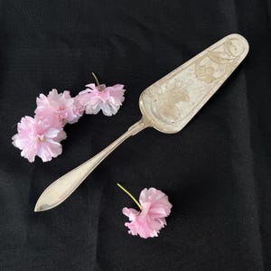 Op de afbeelding: Een zilveren taartschep met een bloemmotief op het blad, naast delicate roze kersenbloesems. De schep heeft een lange steel. De achtergrond is effen zwart.