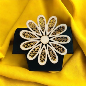 May include: A white and gold flower-shaped brooch with a delicate, intricate design. The brooch is displayed on a black background.